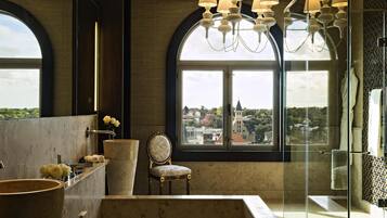 Un baño con una bañera grande, una silla, un tocador con espejo y vistas a un edificio a través de una ventana.