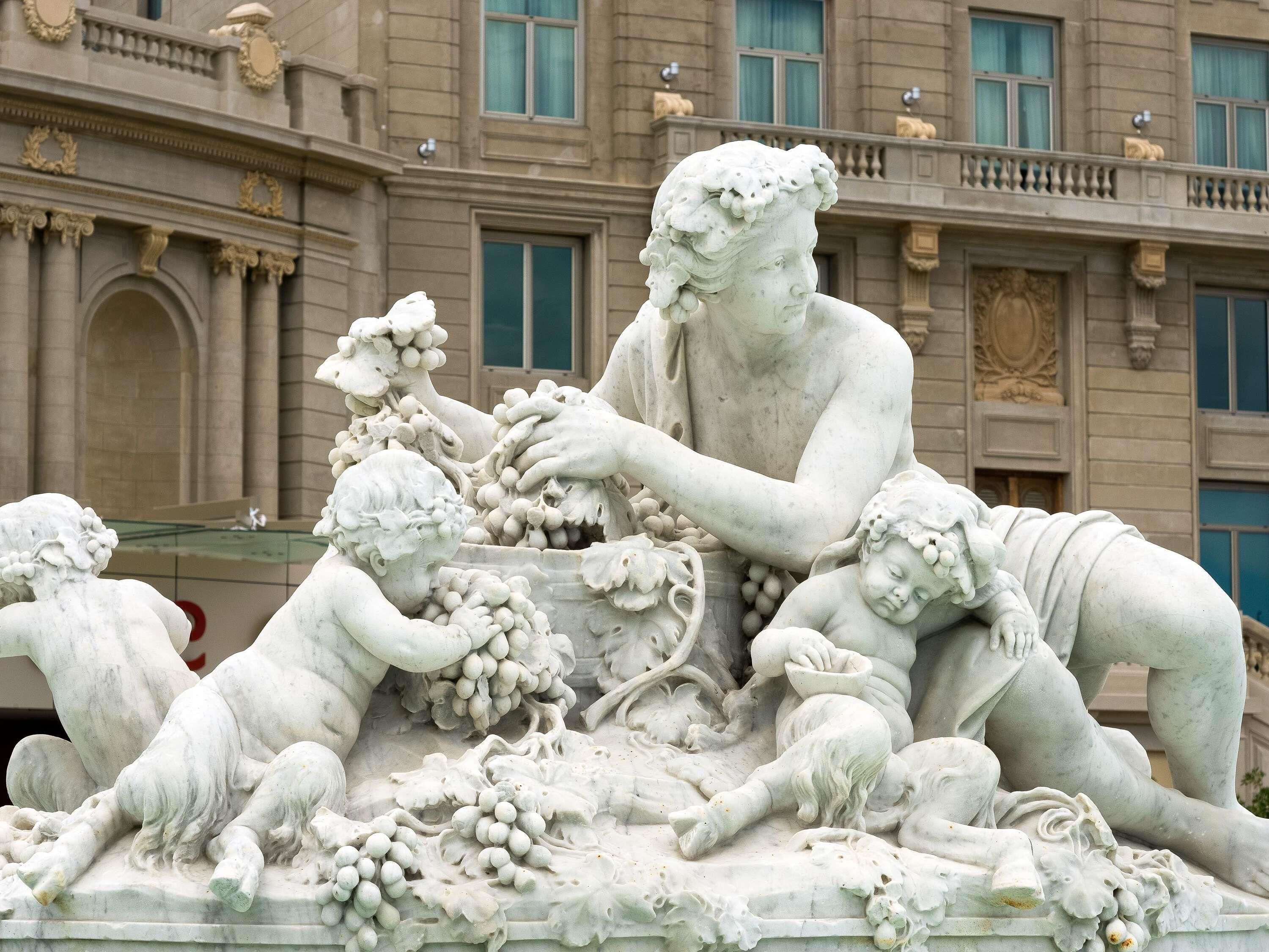 A detailed marble sculpture of a seated figure with children, set against a building facade with classical architectural elements.