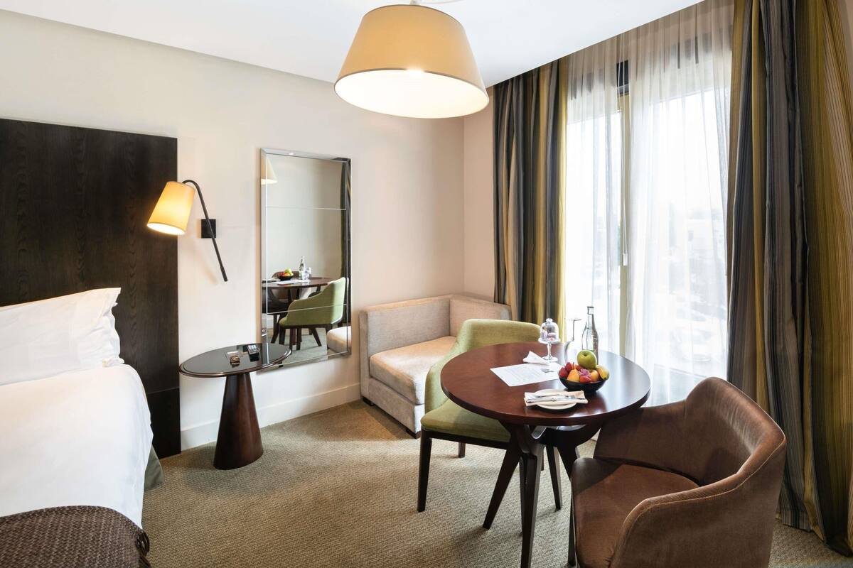 Une chambre d’hôtel comprenant un lit, un bureau, une chaise, un canapé, une table ronde avec un panier de fruits et un grand miroir.