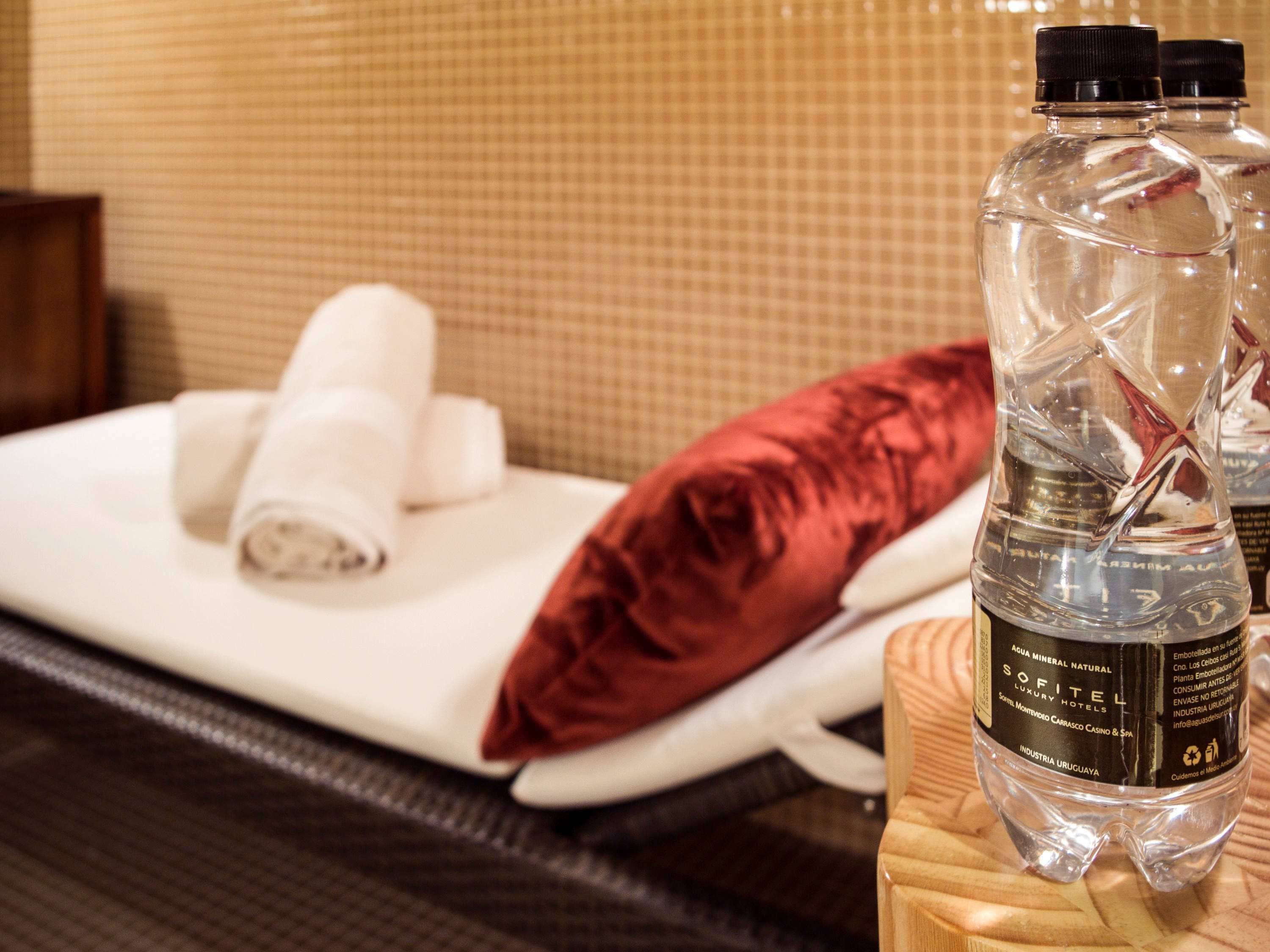Une chambre d’hôtel avec un lit soigneusement fait, un oreiller rouge et deux bouteilles d’eau posées sur une table de chevet en bois.