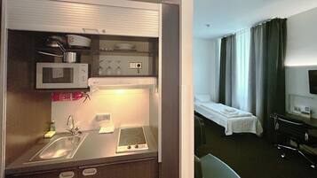 Ein kleines Hotelzimmer mit Küchenzeile, inklusive Mikrowelle, Herd und Spüle. Es gibt ein Bett mit weißer Bettdecke sowie einen kleinen Tisch mit einem Stuhl.