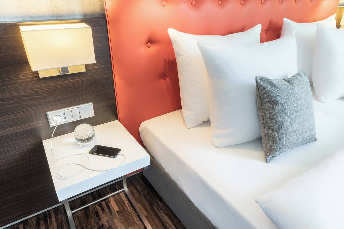 Un lit bien fait, avec des oreillers blancs et gris, une table de chevet avec une lampe, un téléphone et une prise électrique.