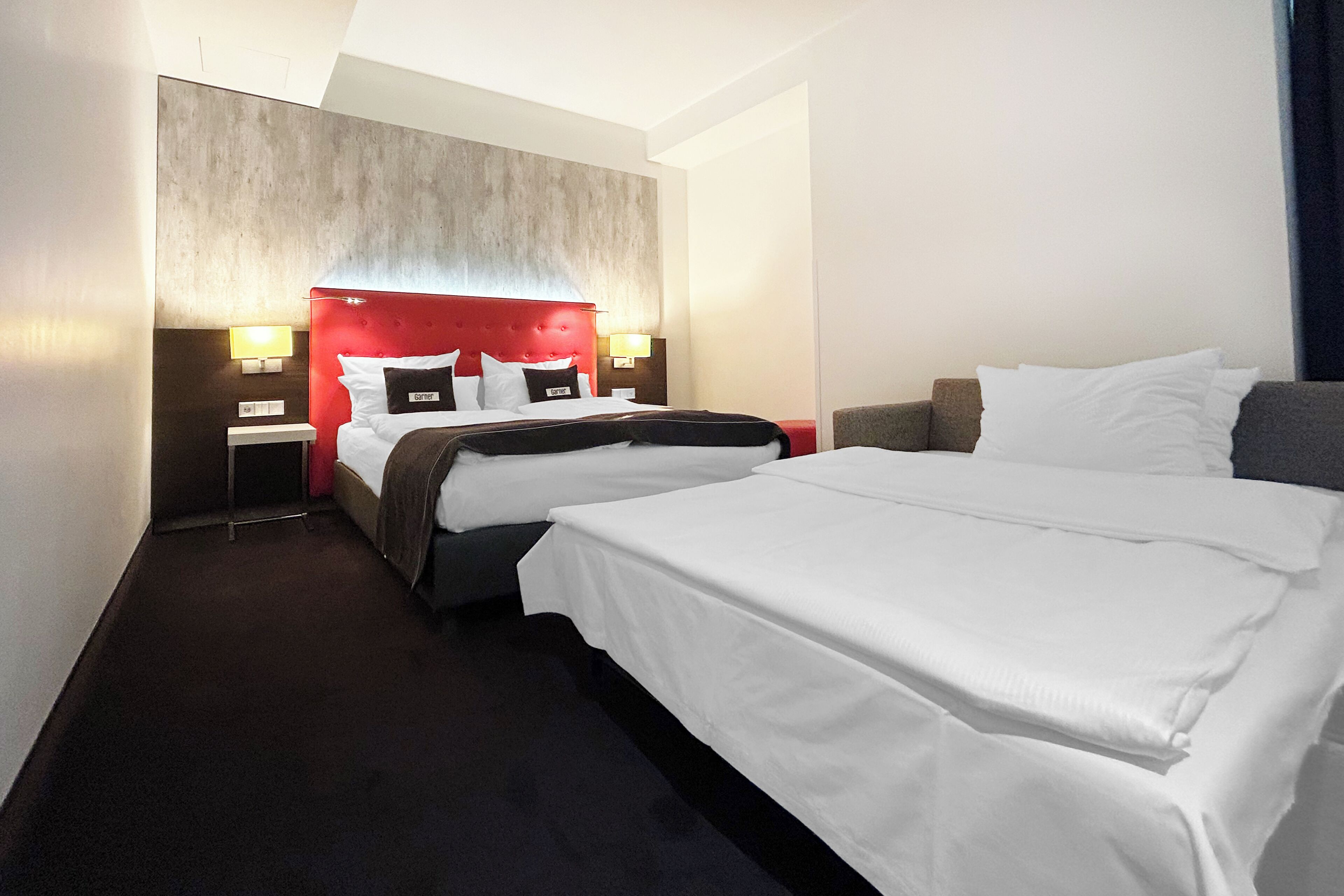 Habitación de hotel con dos camas, cabecero rojo, lámparas de noche y alfombra oscura.
