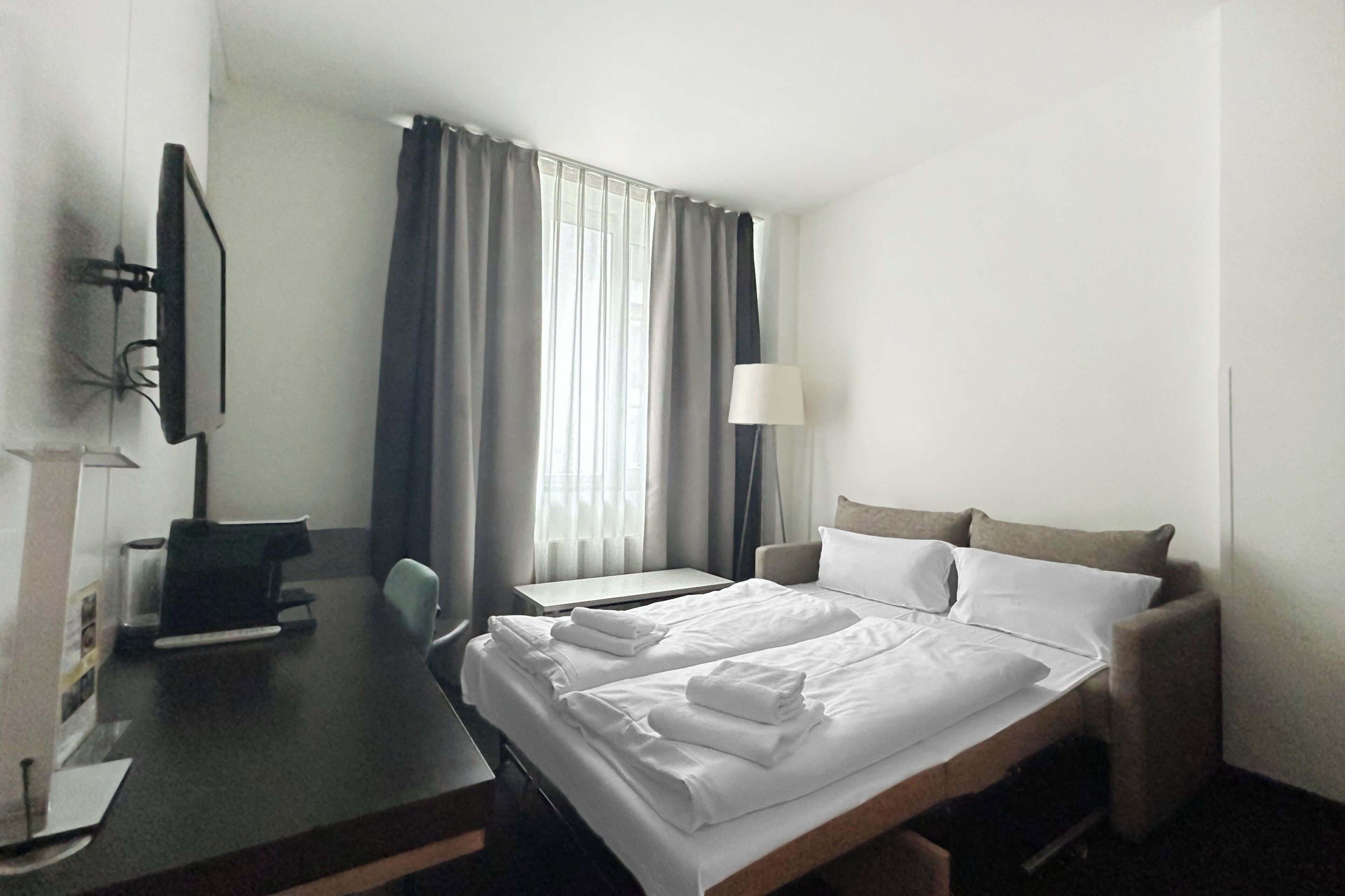 Hotellihuone, jossa on sänky, työpöytä, tuoli, televisio ja lamppu.