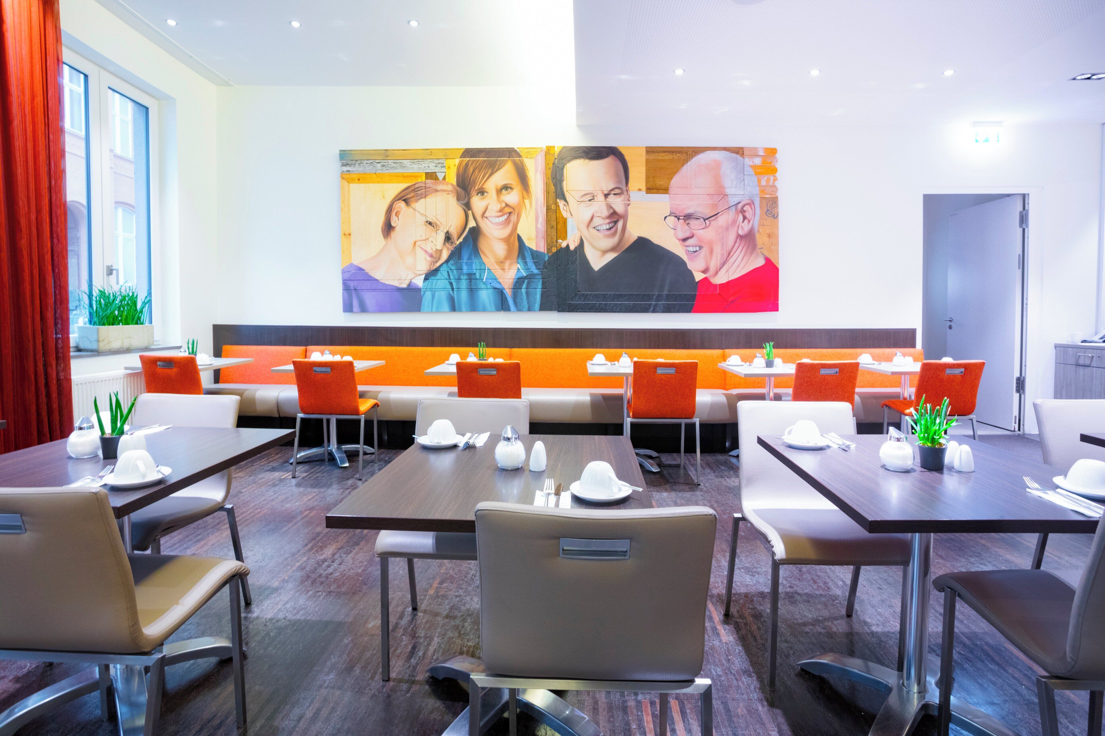 Comedor de hotel con varias mesas y sillas, un mural grande en la pared y una cortina roja a la izquierda.