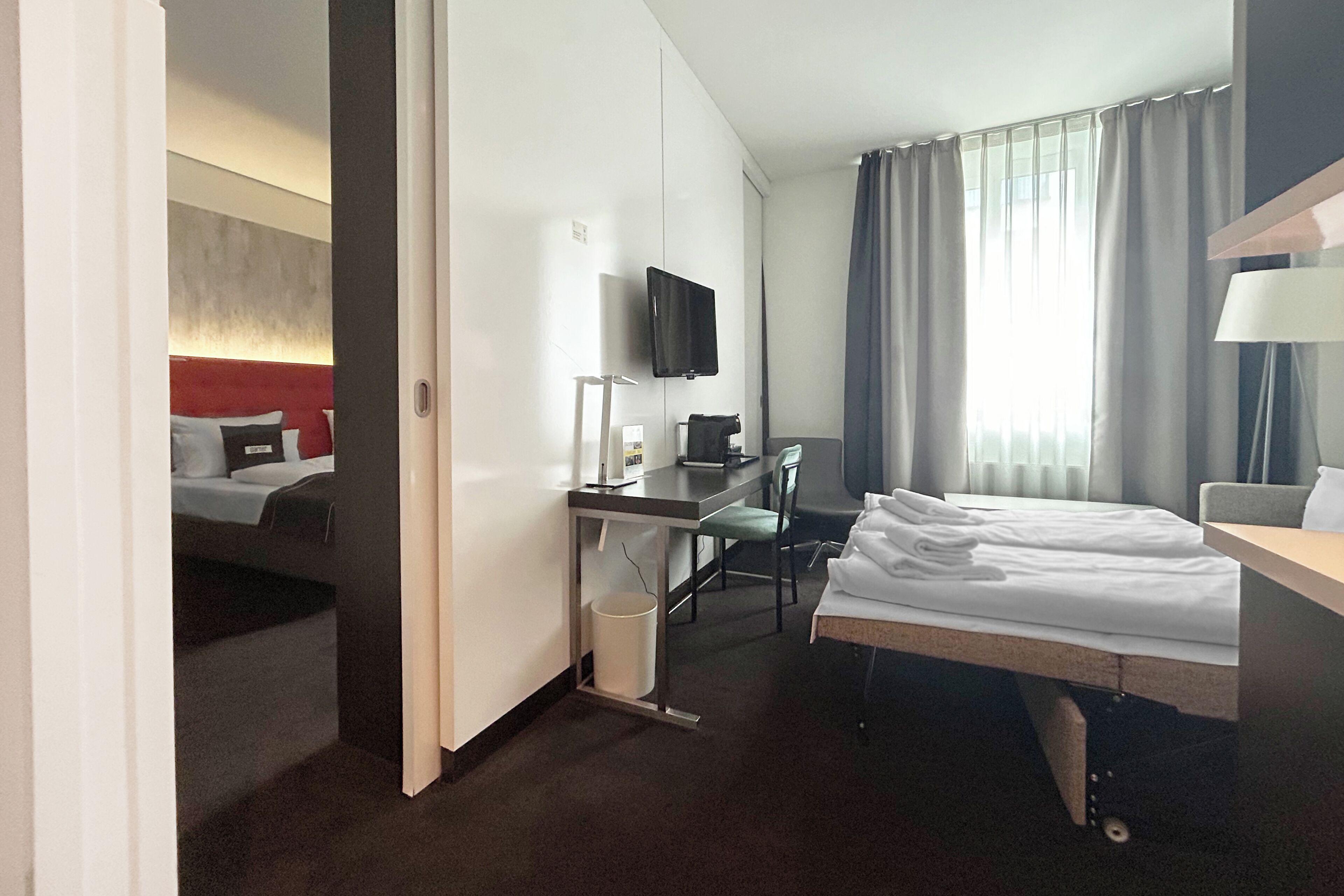 A hotel room with a bed, desk, chair, TV, and a view of the outside.
