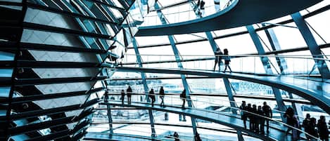 Una estructura arquitectónica moderna con varios niveles y barandas de vidrio, que presenta una escalera en espiral central y personas caminando en los niveles superiores.