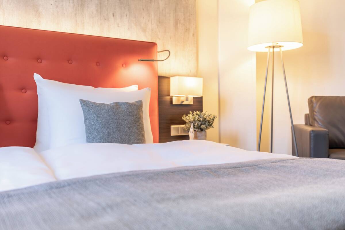 Une chambre d’hôtel avec une tête de lit rouge, un couvre-lit blanc et un oreiller gris. On y trouve également une lampe sur pied et une chaise.