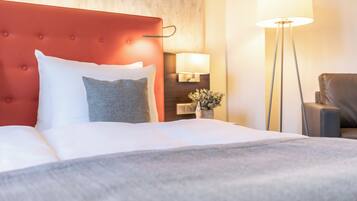Habitación de hotel con cabecera tapizada en rojo, cubrecama blanco y almohada gris. Hay una lámpara de pie y una silla en la habitación.