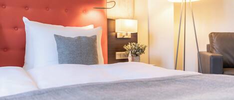 A hotel room with a red upholstered headboard, a white bedspread, and a grey pillow. There is a floor lamp and a chair in the room.