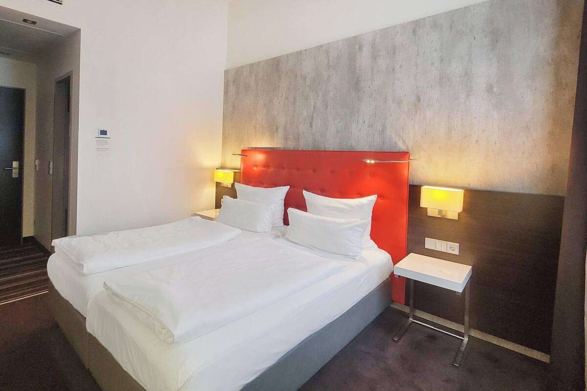 Un lit double avec du linge de lit blanc, une tête de lit rouge, deux lampes de chevet et une petite table de chevet.