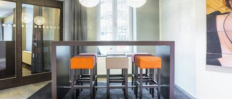 A modern dining area with a dark wooden table, three bar stools with orange and beige cushions, and two crystal chandeliers.