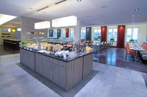 Un'area ristorazione moderna in hotel con un'isola buffet centrale, diverse disposizioni di posti a sedere e ampie finestre con vista.