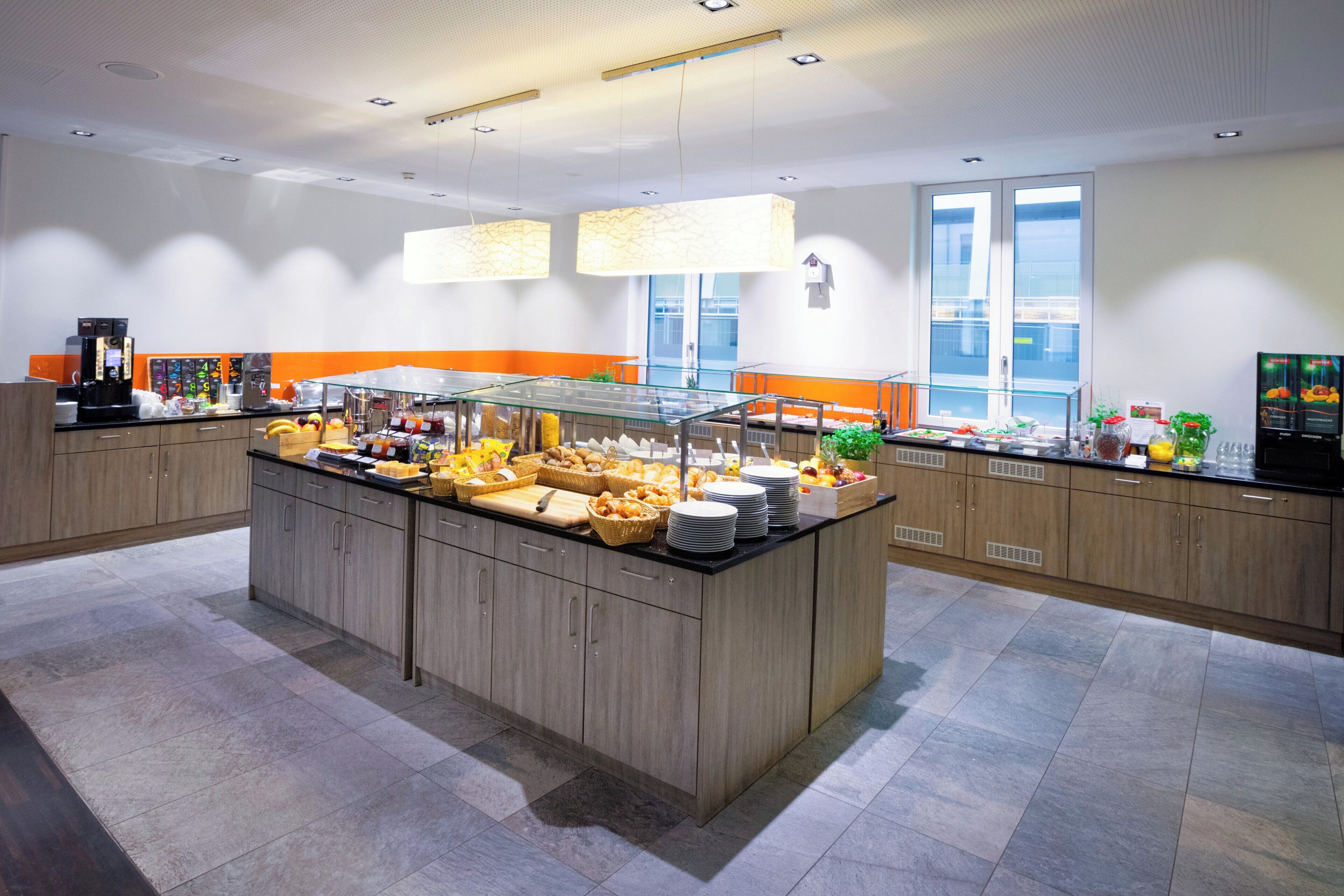 Un área de desayuno moderno en un hotel, con una isla central de buffet, muebles de madera y un amplio ventanal que proporciona luz natural.
