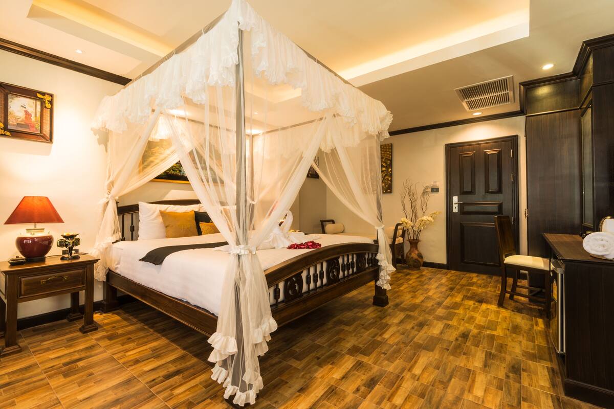 A four-poster canopy bed with a wooden frame and white bedding, a nightstand with a red lamp, and a dark wooden wardrobe.