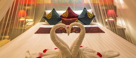 Une chambre d’hôtel avec un lit agrémenté d’une décoration en forme de cœur réalisée avec des serviettes et des pétales de rose, une tête de lit aux motifs rouges et dorés, et un mur décoratif orné d’un motif floral.
