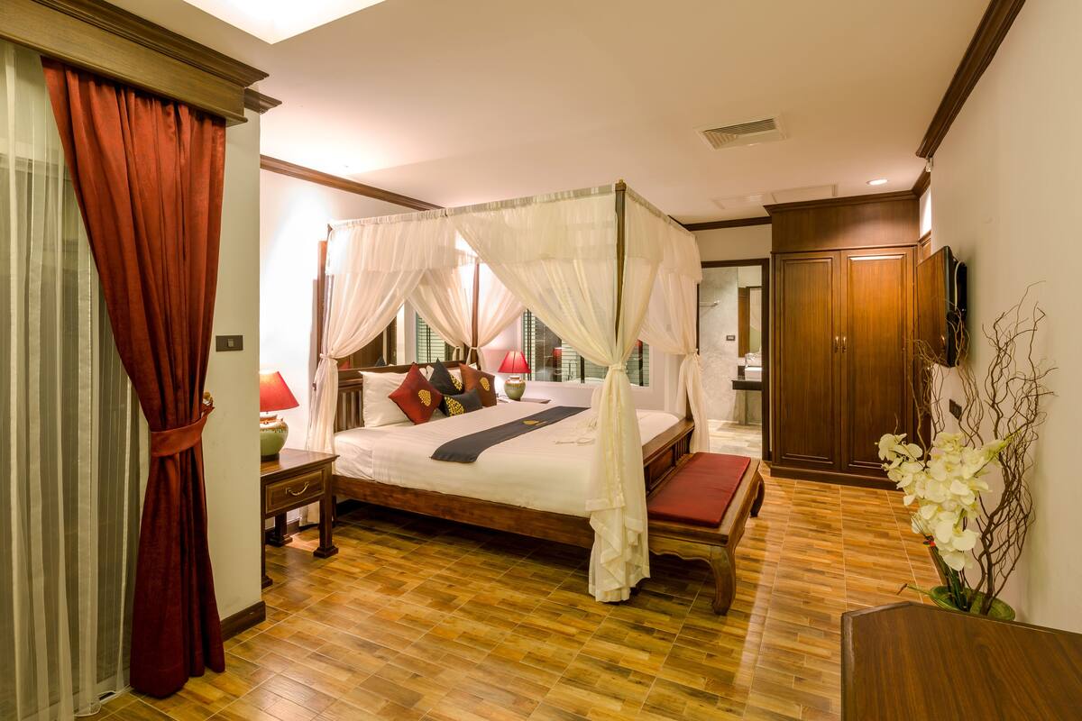 A four-poster canopy bed with a wooden frame, a nightstand with a lamp, and a red curtain.