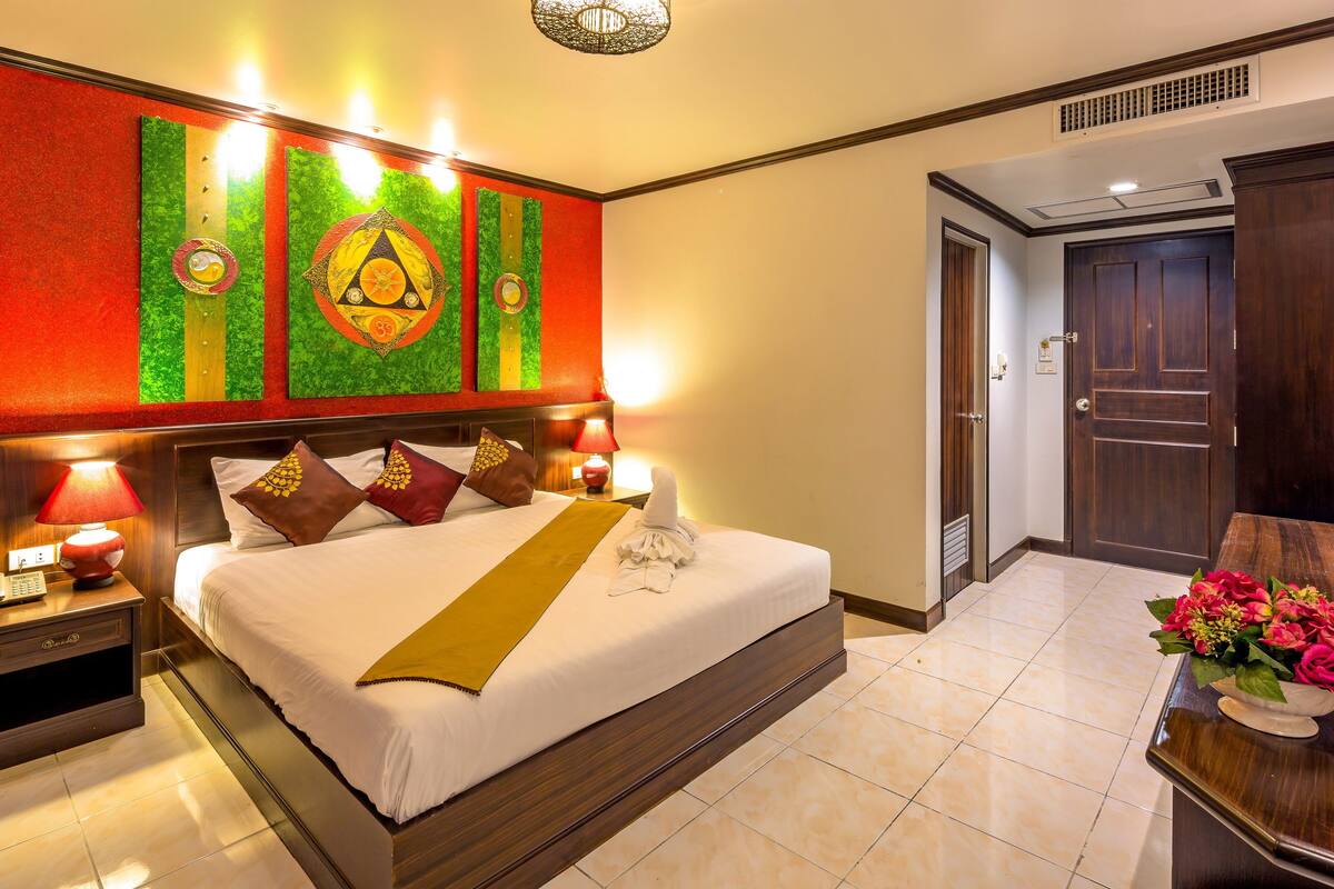 A hotel room with a large bed, a headboard with artwork, a nightstand with a lamp, and a desk with a flower arrangement.
