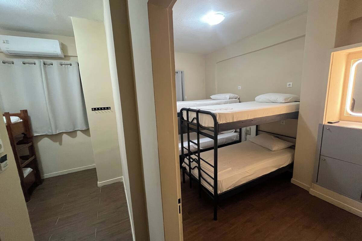 A dormitory room with bunk beds, a wooden ladder, a white air conditioning unit, and a small window with a light fixture.