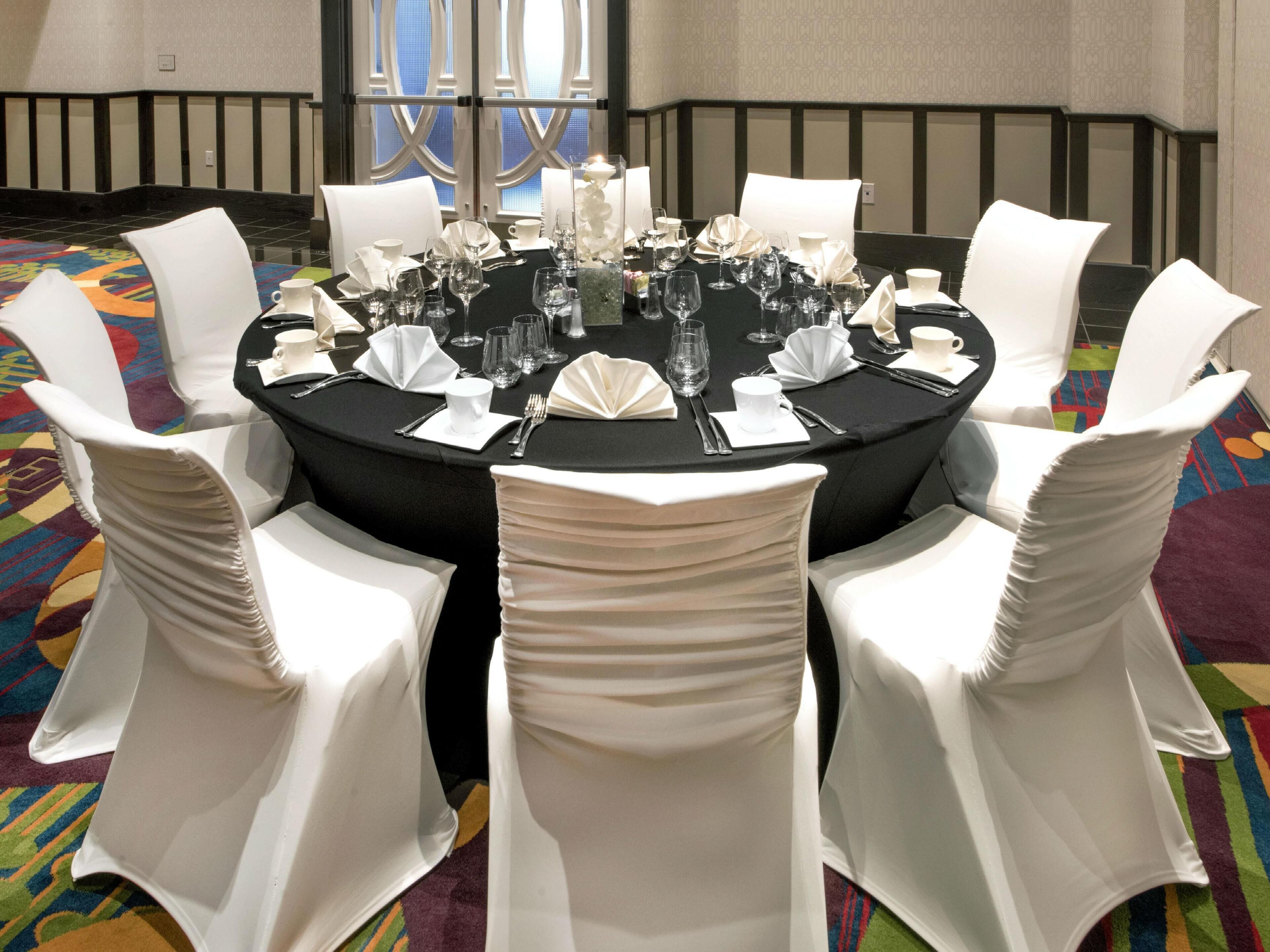 Mesa redonda dispuesta para una comida, con sillas blancas, mantel negro y un centro de mesa con una vela.