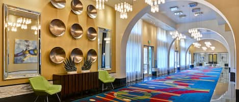 A hotel lobby with a long, colorful carpet, green chairs, a large mirror, and modern chandeliers.
