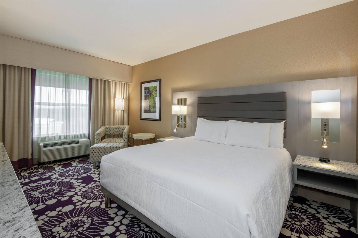 A hotel room with a large bed, a chair, a desk, and a wall-mounted TV.