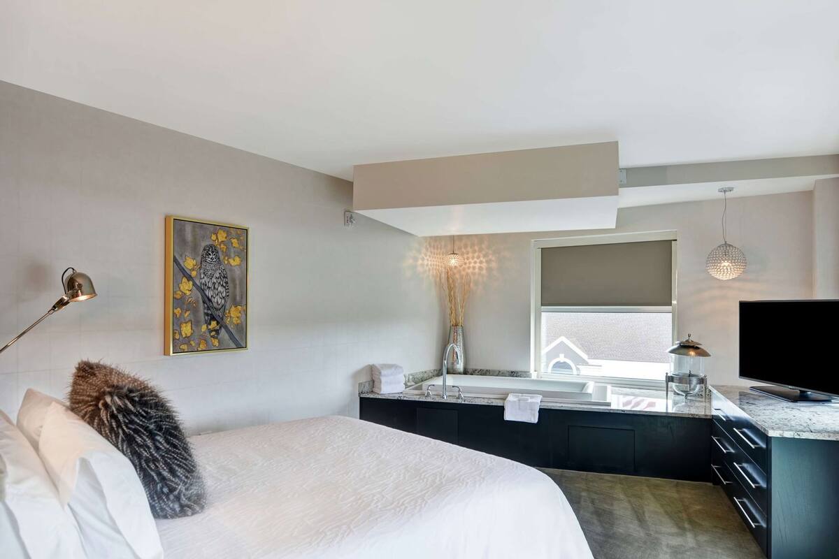 A modern bedroom with a bed, a desk, a television, a painting, and a window.