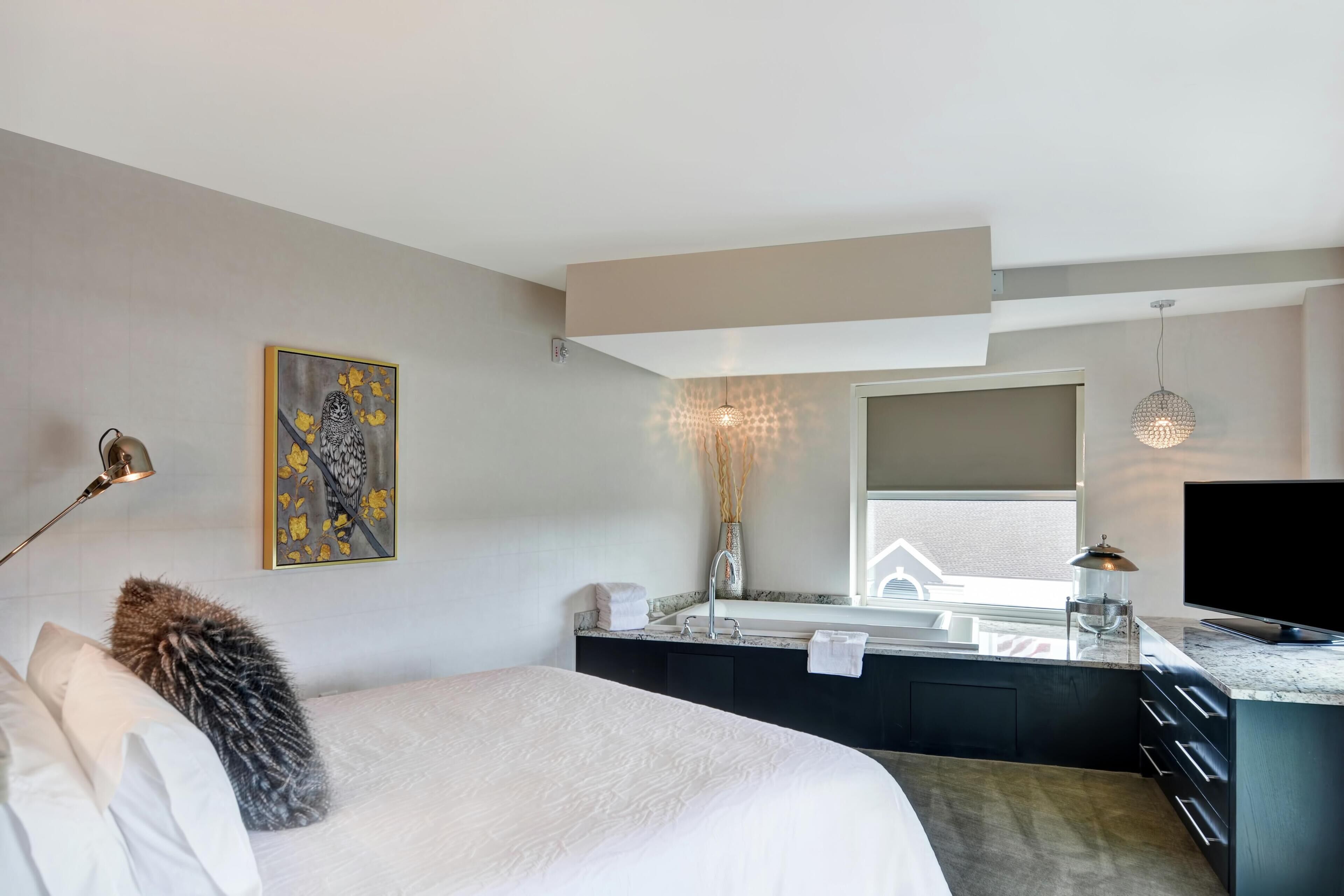 A modern hotel room with a bed, a desk area with a sink, a flat-screen TV, and a painting on the wall.