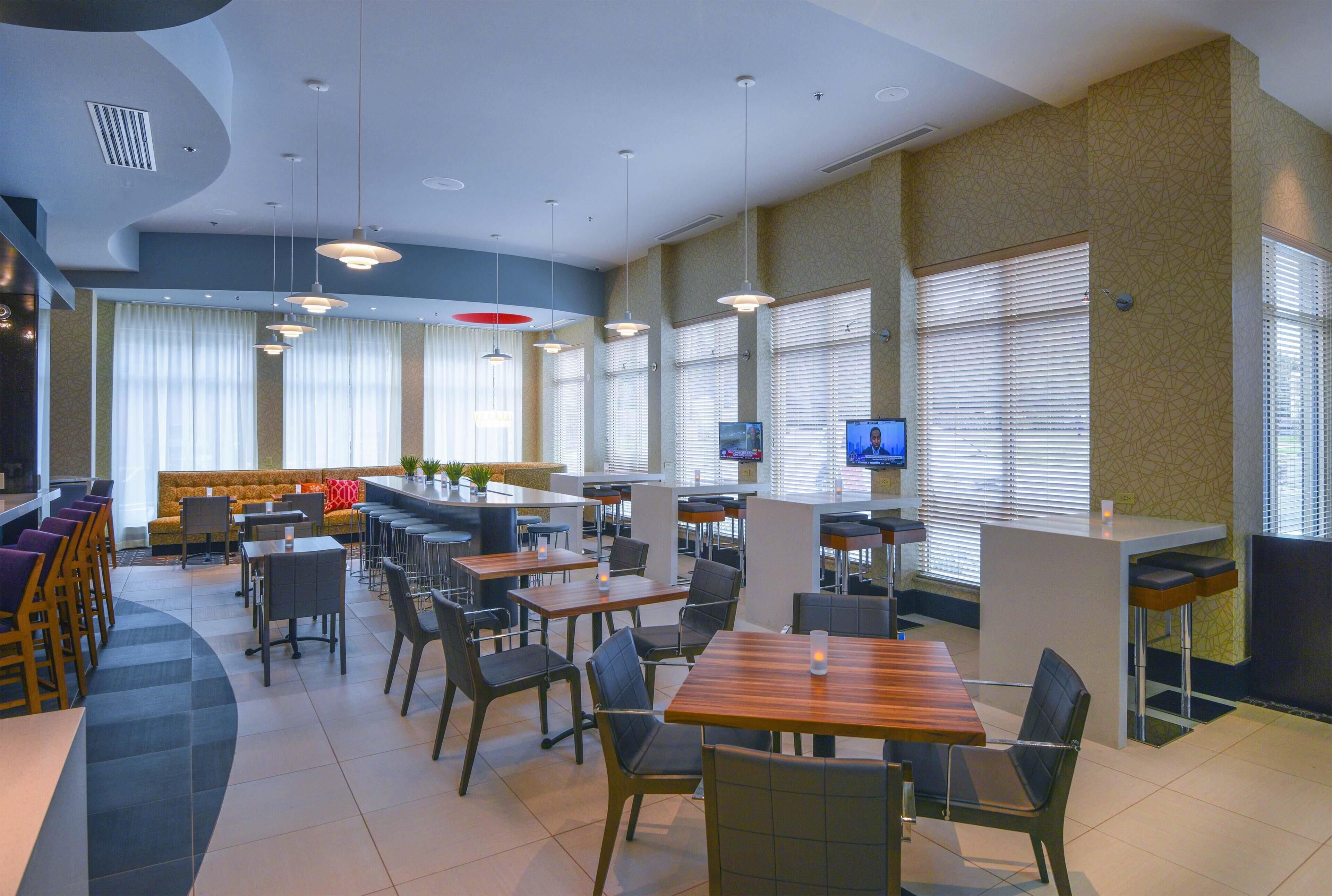 A modern restaurant with high tables and chairs, a bar area, and large windows with blinds.