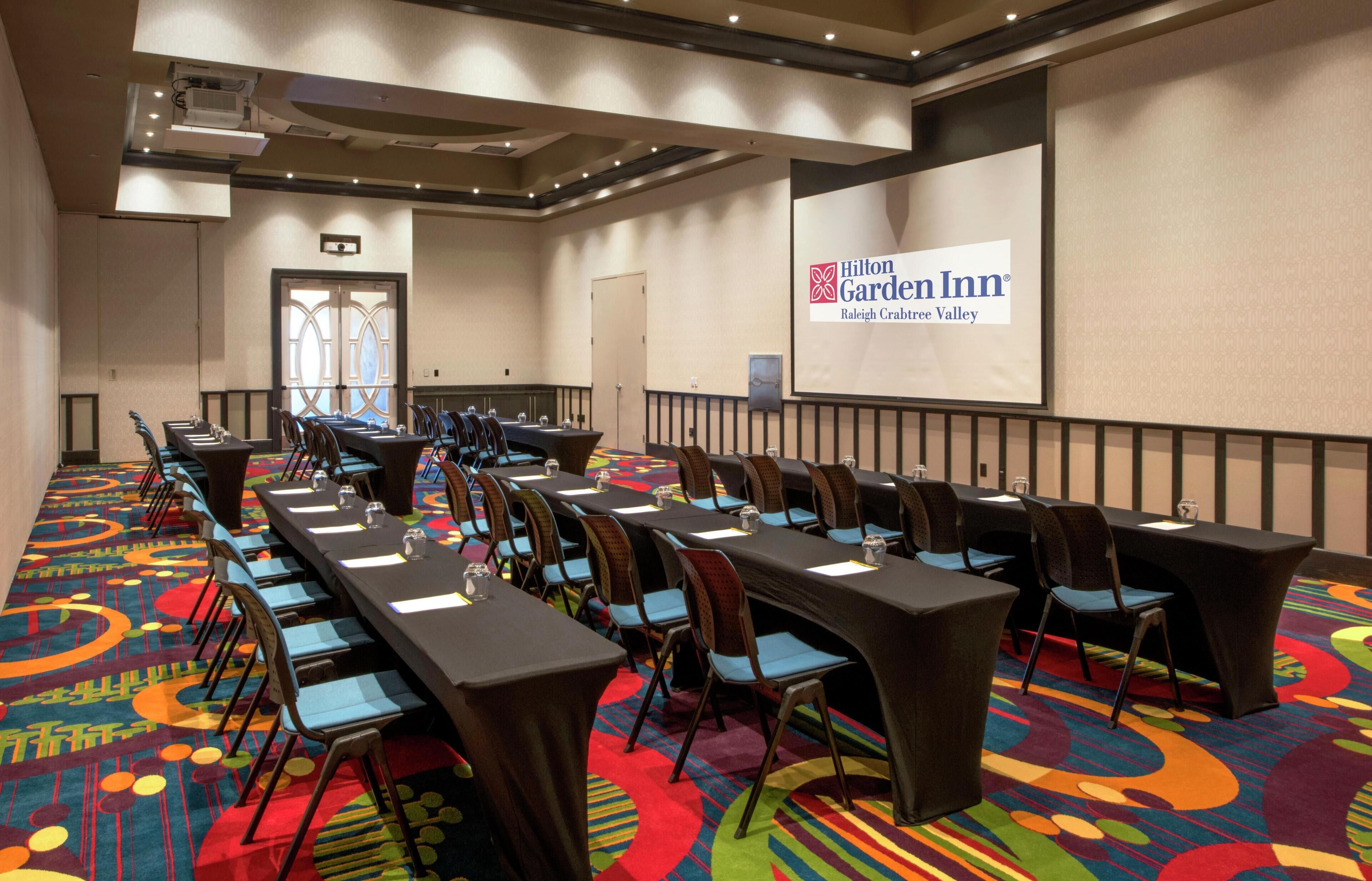Ein Konferenzraum im Hilton Garden Inn mit langen Tischen, Stühlen und einer Projektionsleinwand, die den Namen des Hotels zeigt.