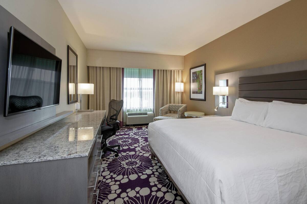 A hotel room with a large bed, a desk, a chair, a TV, and a window with curtains.