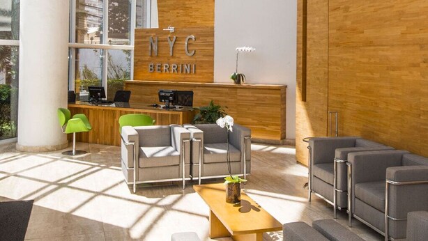 Una moderna zona de recepción con un mostrador de madera que dice "NYC BERRINI", sofás grises y una máquina de café.