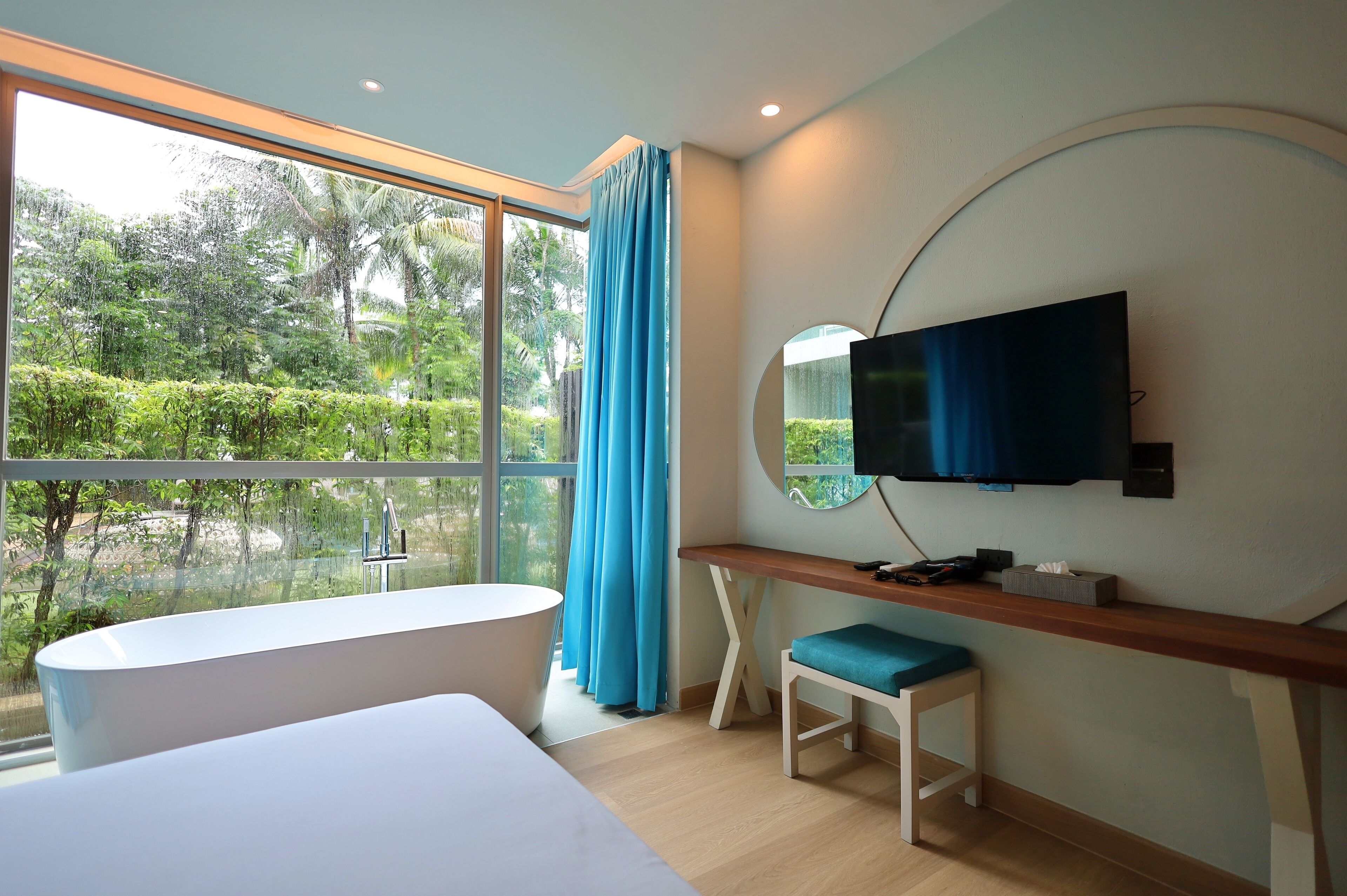 A modern hotel room with a large window, a bathtub, a desk, and a television.