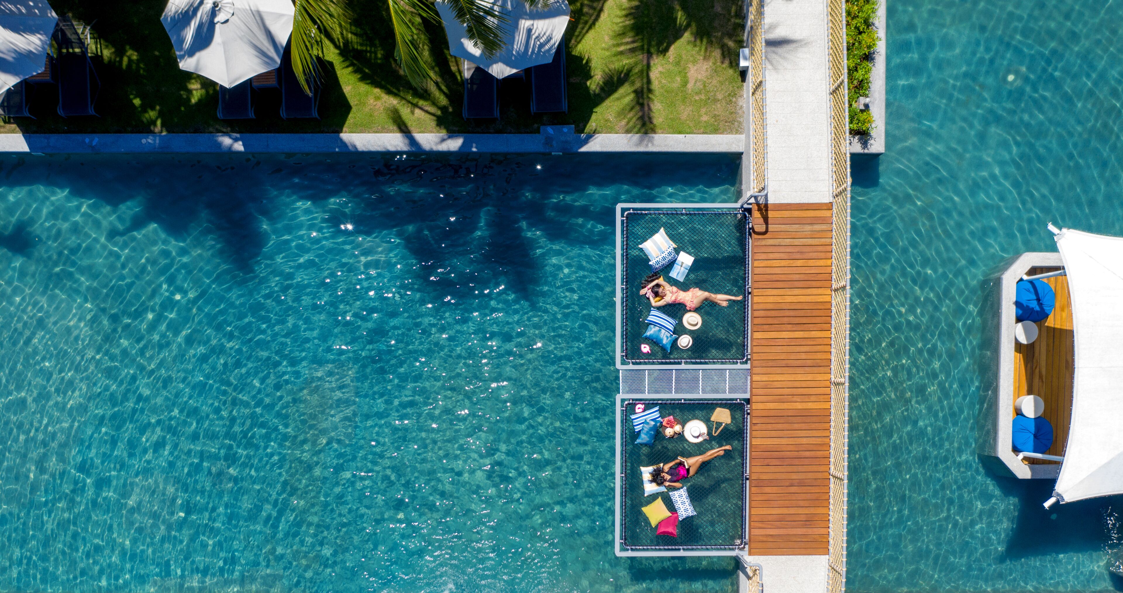 Vue aérienne d’un complexe hôtelier avec une piscine d’eau cristalline, une terrasse en bois et des chaises longues.
