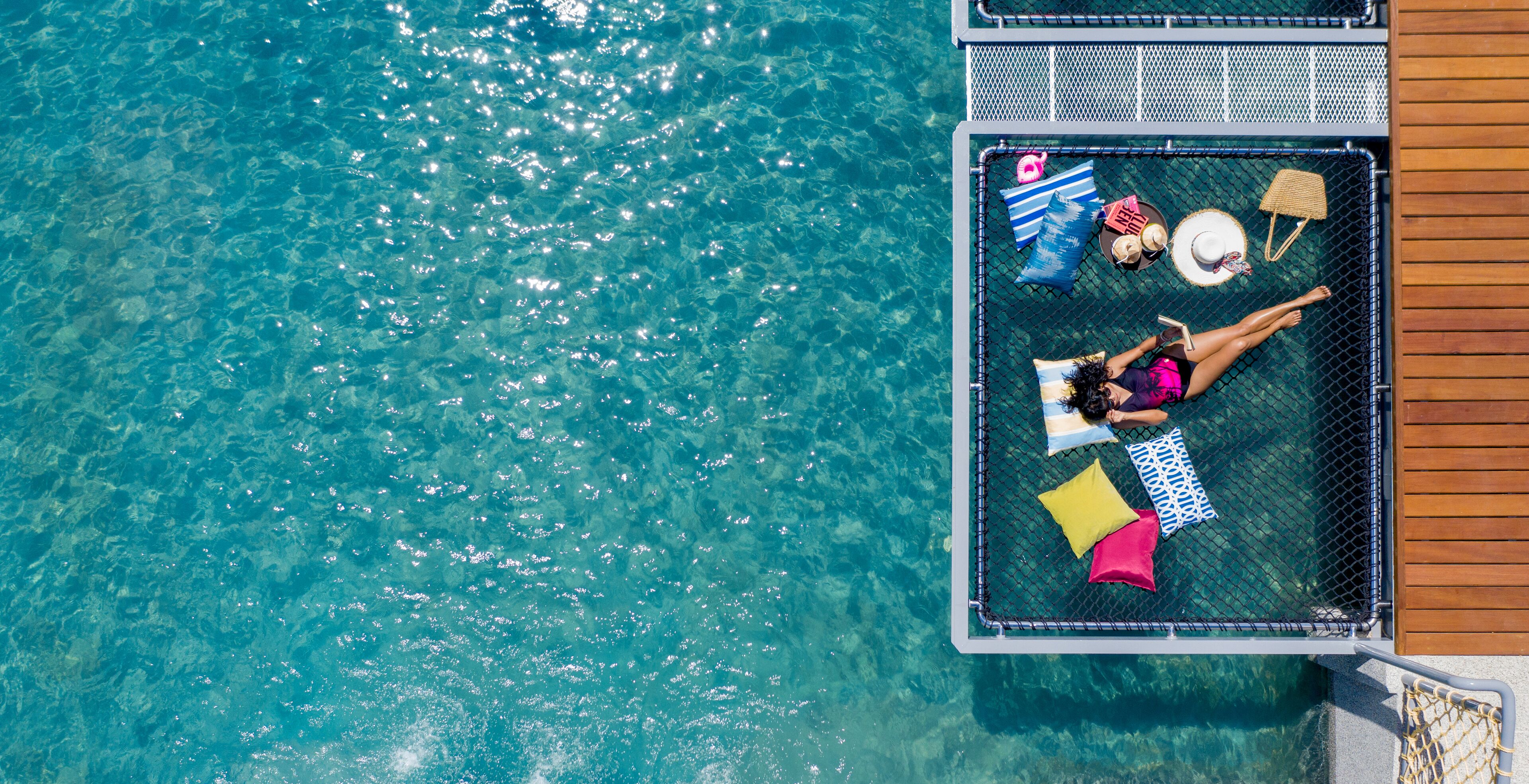 A floating platform with a hammock and colorful pillows, surrounded by clear blue water.