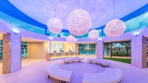 Un espace intérieur moderne doté d’un espace salon circulaire, de grandes lampes sphériques décoratives et d’un comptoir d’accueil offrant une vue sur une piscine extérieure.