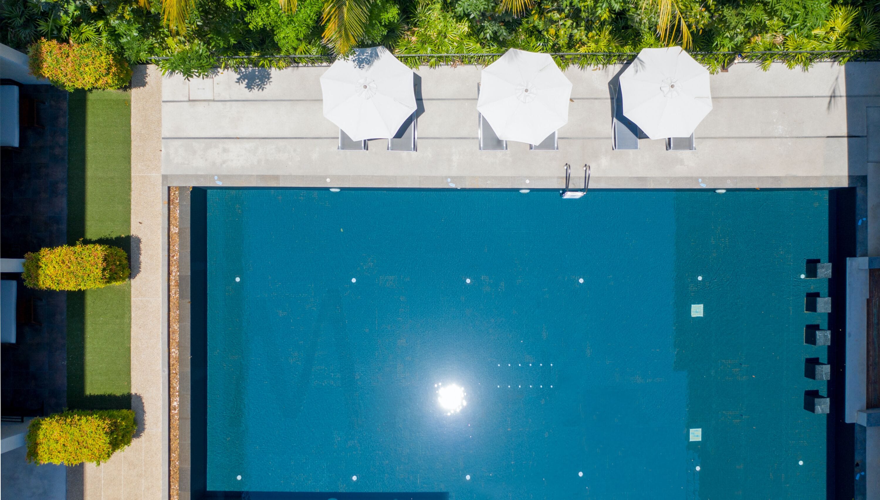 Vista aérea de uma piscina retangular com três guarda-sóis brancos e um deck revestido de azulejos ao redor.