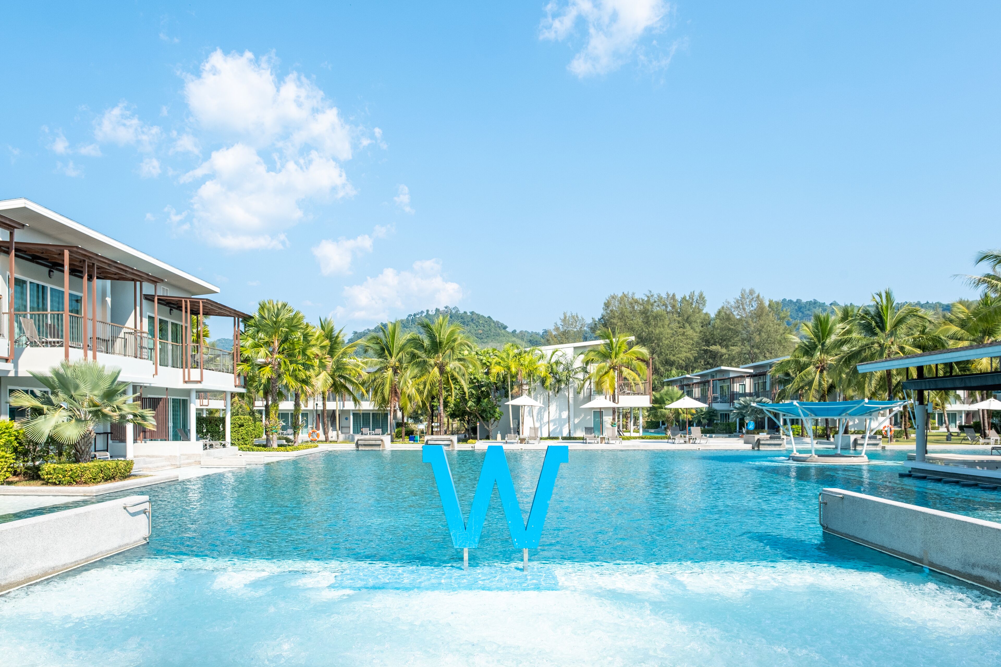 A swimming pool with a large blue 'W' sculpture in the center, surrounded by resort buildings and palm trees.