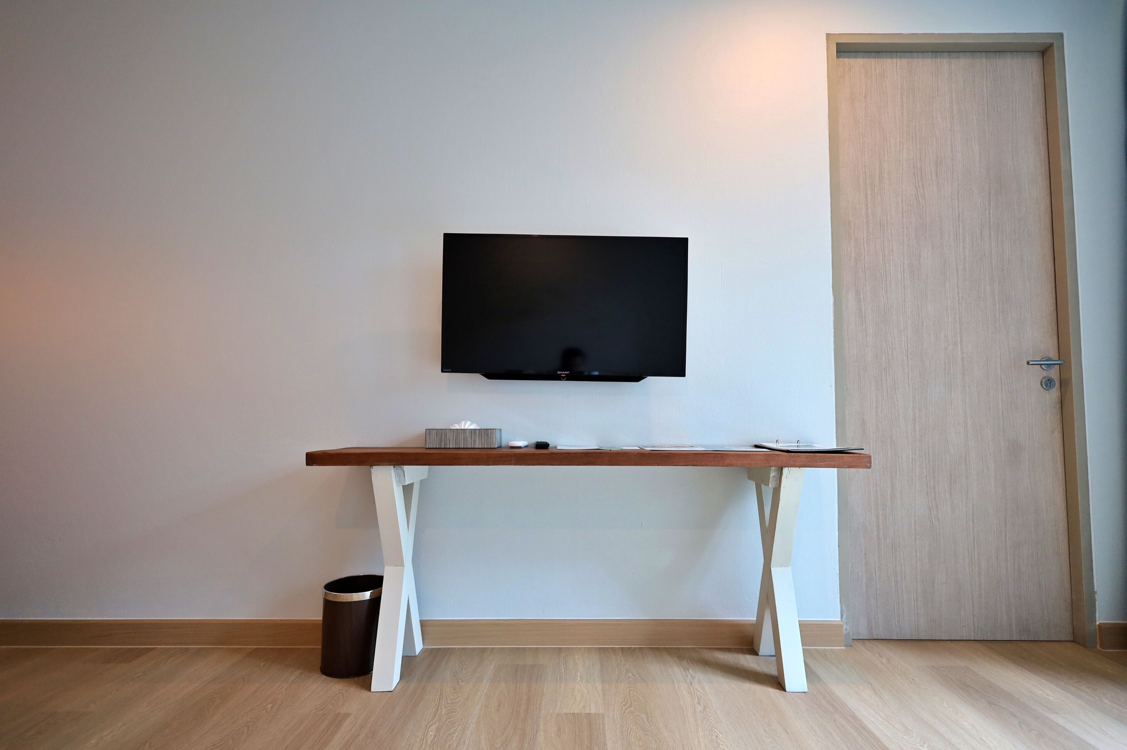 Une pièce avec un téléviseur fixé au mur, une table en bois et une poubelle à côté, ainsi qu’une porte en bois.