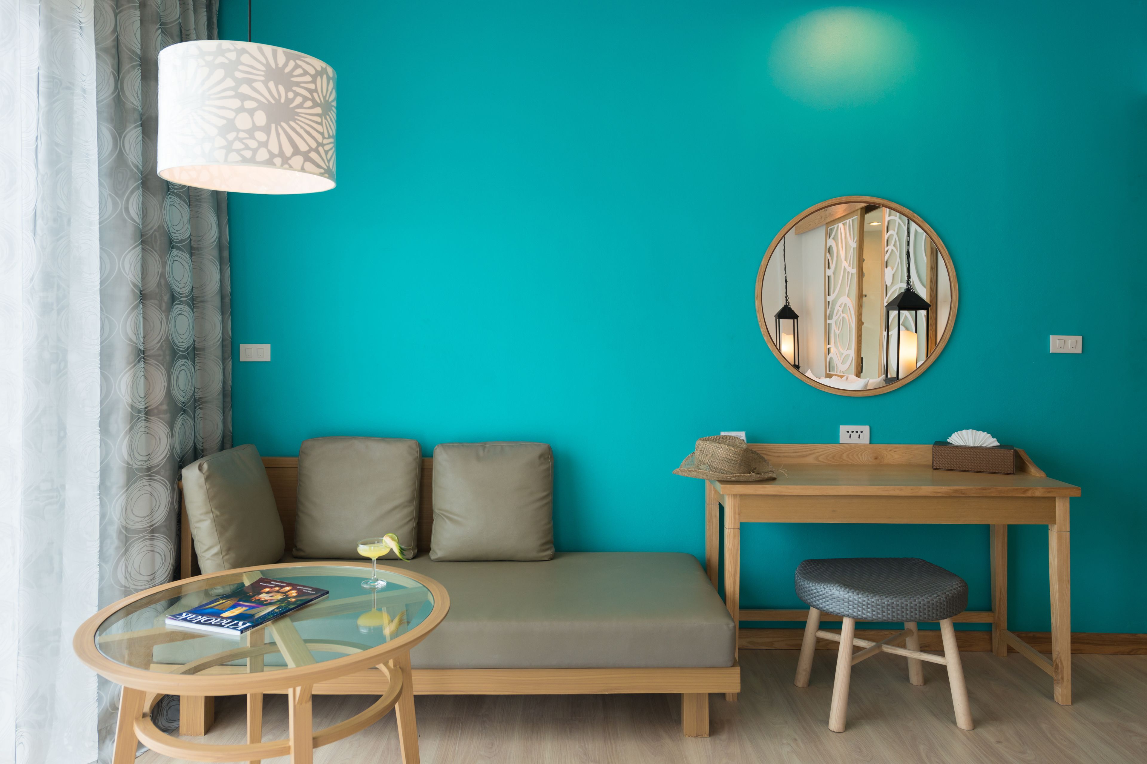 A modern room with a teal wall, a round mirror, a wooden desk, a cushioned bench, and a glass table.