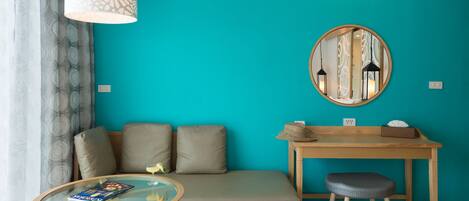 A modern room with a teal wall, a round mirror, a wooden desk, a cushioned bench, and a glass table.
