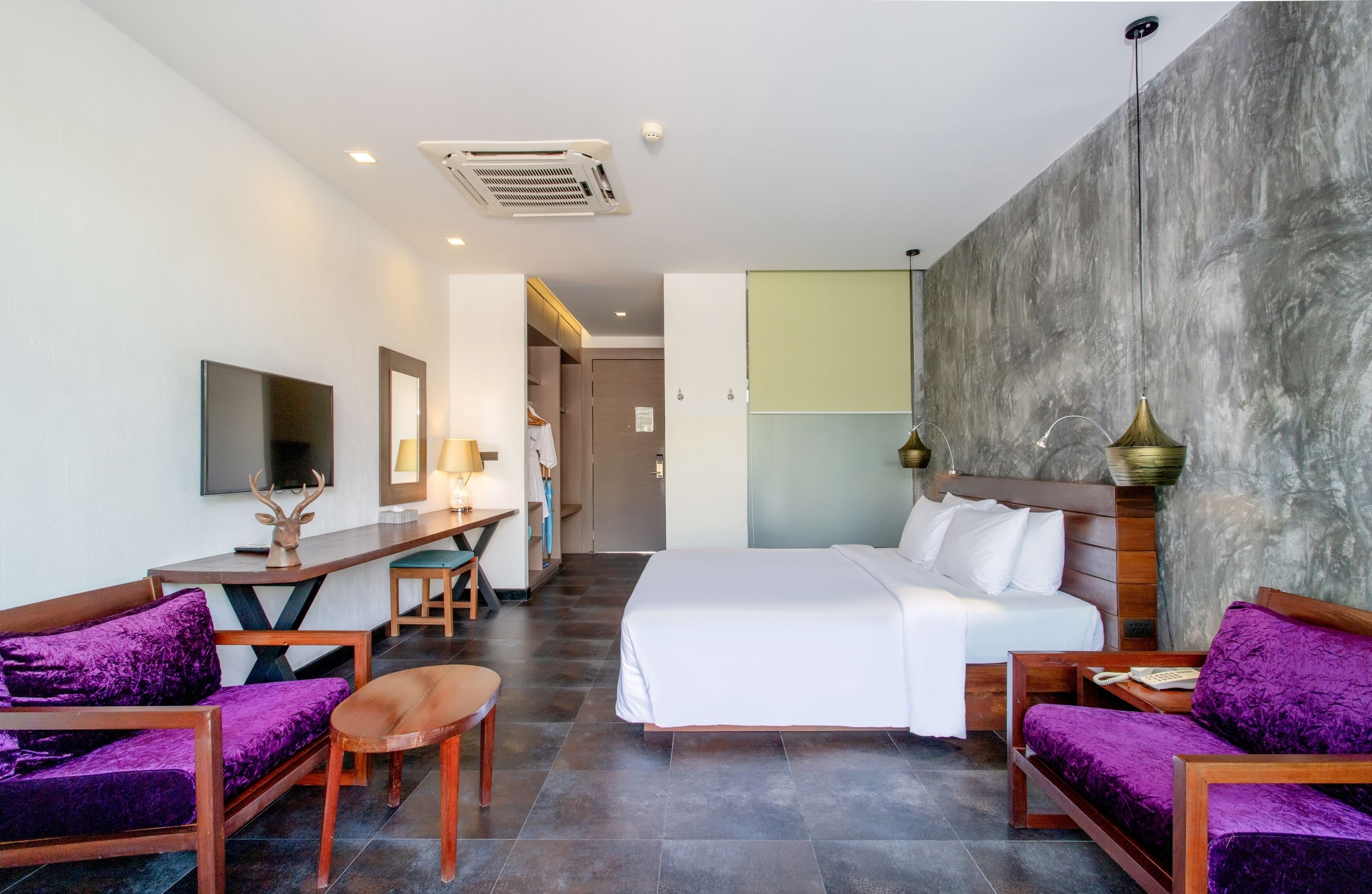 Habitación de hotel con cama, dos sofás morados, un escritorio de madera, televisión y una gran pared de piedra.