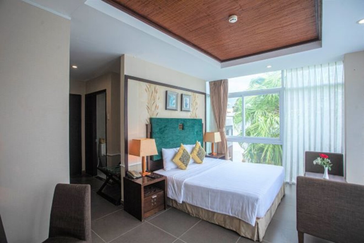 A hotel room with a large bed, two bedside tables with lamps, a chair, and a view of greenery through the window.