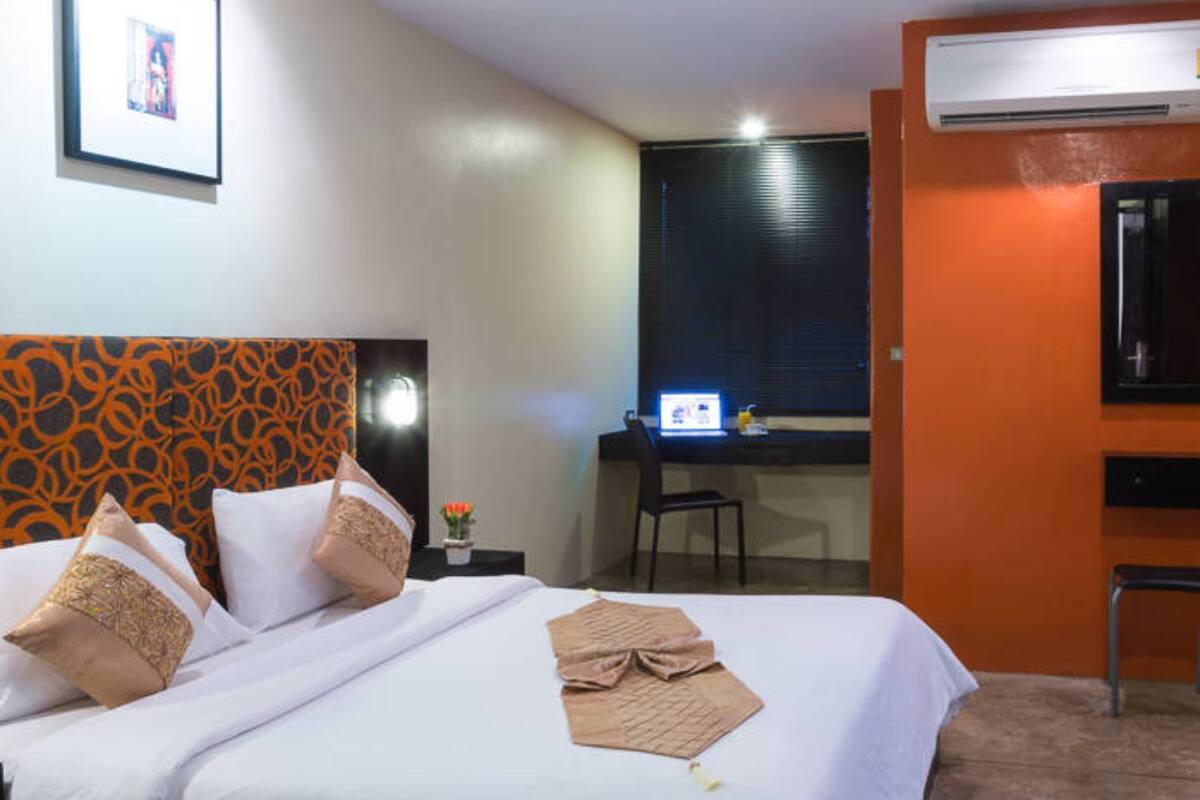 A hotel room with a bed, a desk with a computer, an air conditioner, and a vase.