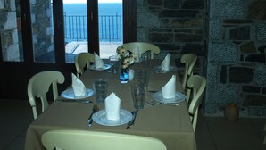 A dining table set for six with white chairs, a stone wall, and a view of the sea through a large window.