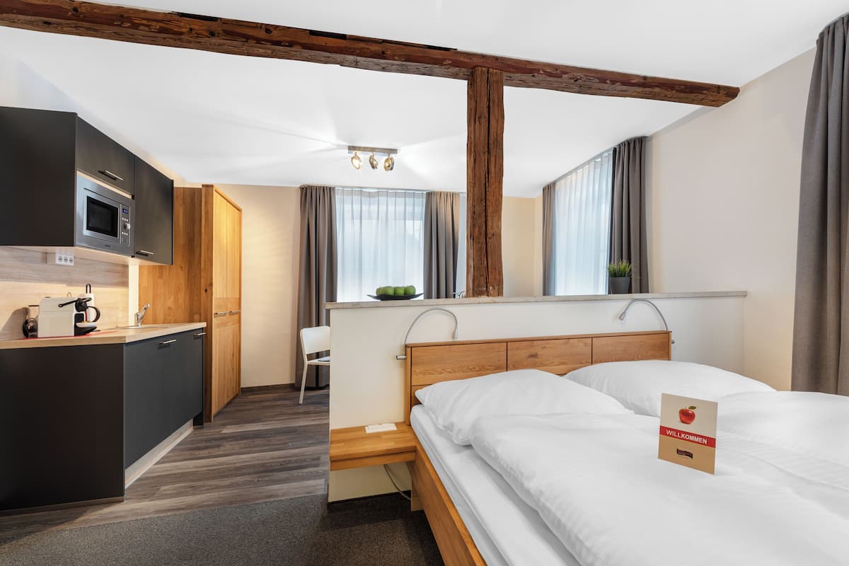 A modern hotel room with a wooden ceiling, a bed, a desk, a kitchenette, and a view of the outdoors.