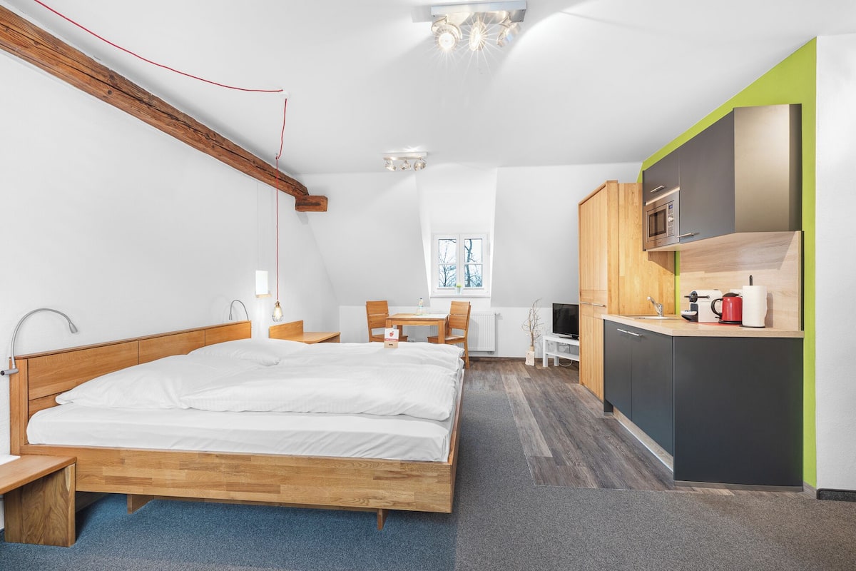 A bedroom with a wooden bed, a small desk, a chair, a window, and a kitchenette with a microwave and toaster.