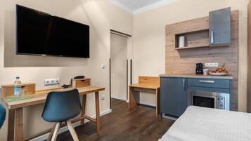 A compact hotel room with a bed, a desk with a chair, a television, a kitchenette, and a small bathroom.