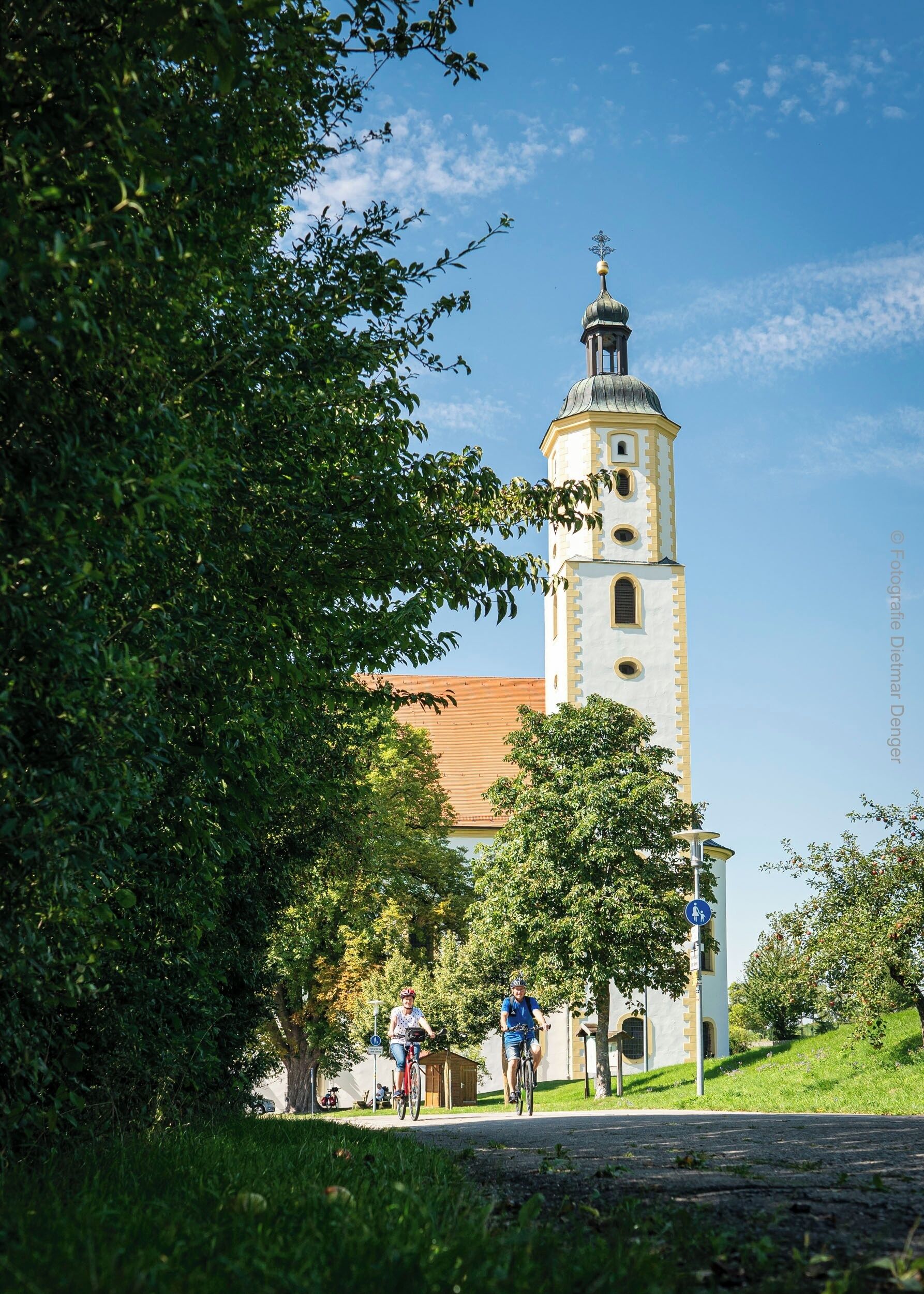 A church with a bell tower, surrounded by greenery and a paved path.