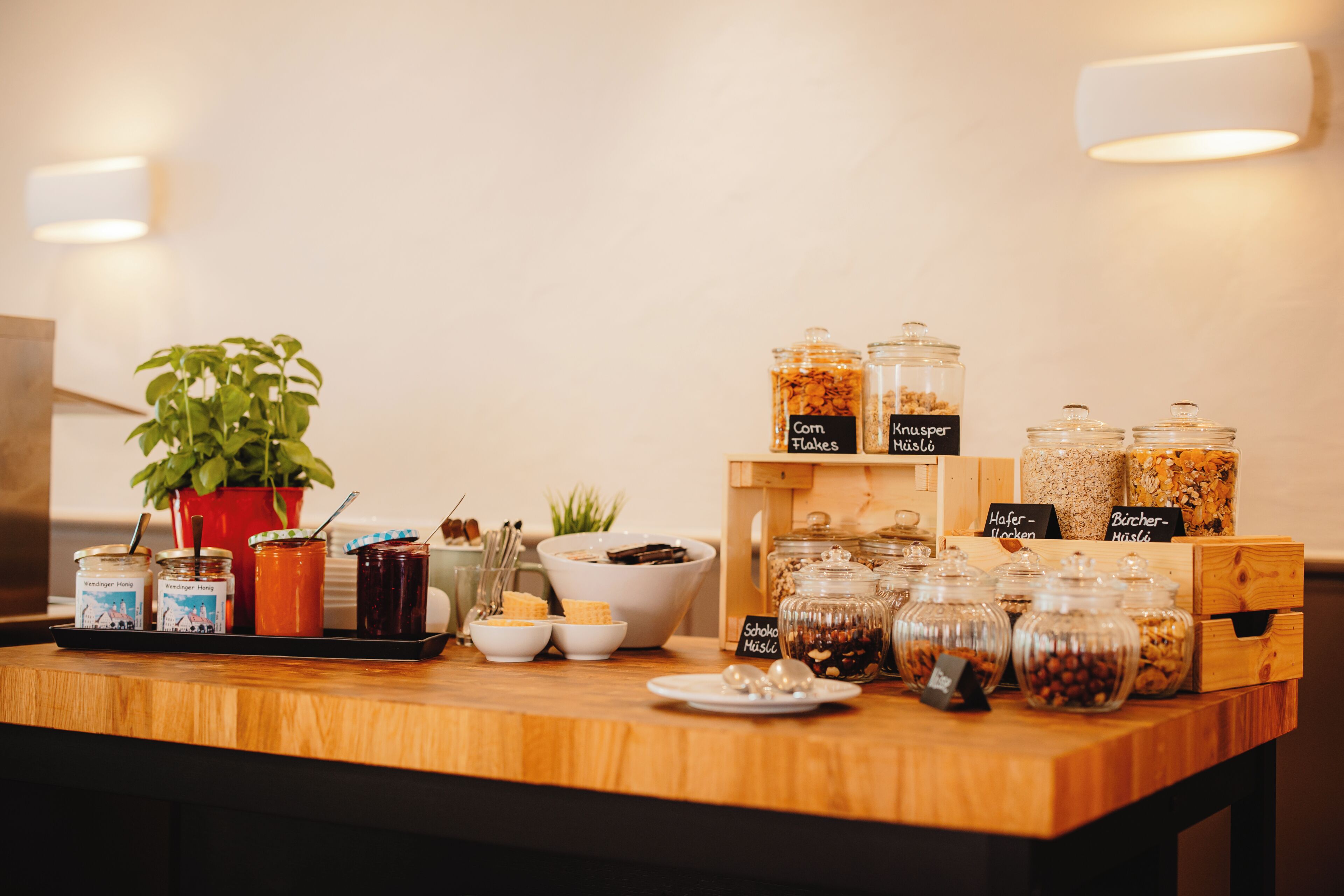 Un buffet proposant divers aliments présentés dans des récipients étiquetés avec des noms tels que « Corn Flakes », « Knusper » et « Hafer-Mochen ».